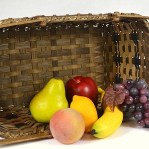 May include: A woven basket filled with fruit, including a red apple, a yellow pear, a yellow peach, a yellow lemon, a yellow banana, and a bunch of purple grapes.