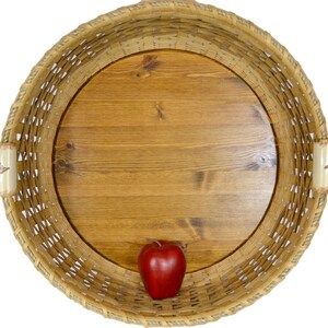 Pu&ograve; includere: Un cesto rotondo intrecciato con una base in legno e una mela rossa all'interno. Il cesto &egrave; fatto di materiali naturali e ha un aspetto rustico.