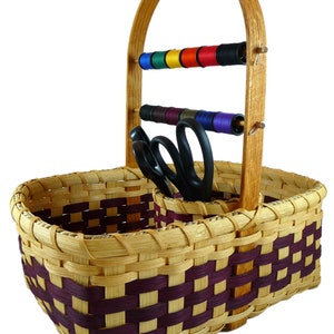 May include: A woven basket with a wooden handle and two compartments. The basket has a purple and natural woven pattern. The handle has two wooden dowels with spools of thread on them. There are also two pairs of scissors in the basket.