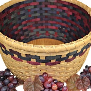 May include: A woven basket with a natural brown base and a multi-colored design of red, blue, and black. The basket is empty and surrounded by grapes.