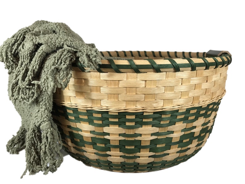 May include: A large, woven basket with a green and natural colored pattern. The basket has a handle and is filled with a soft, green blanket.
