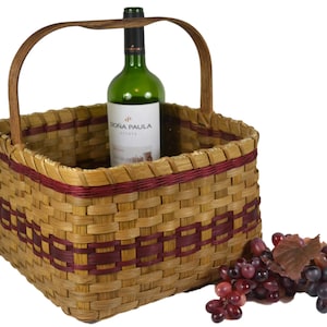May include: A woven basket with a wooden handle, featuring a red and brown pattern. The basket contains a bottle of wine and is next to a bunch of grapes.