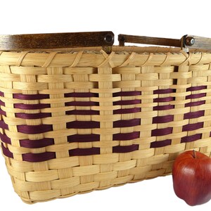 May include: A woven basket with a wooden handle and burgundy accents. The basket is made of natural materials and has a rustic look. An apple is sitting next to the basket.