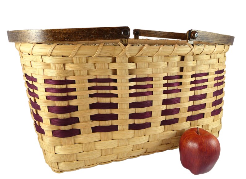May include: A woven basket with a wooden handle and burgundy accents. The basket is made of natural materials and has a rustic look. An apple is sitting next to the basket.