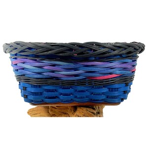 May include: A woven basket with a blue, purple, and black design. The basket has a wooden base and is oval shaped.