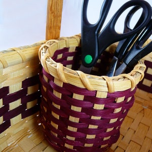 May include: A woven basket with a burgundy and natural colored pattern holds three pairs of black scissors. The basket is sitting on a wooden surface.