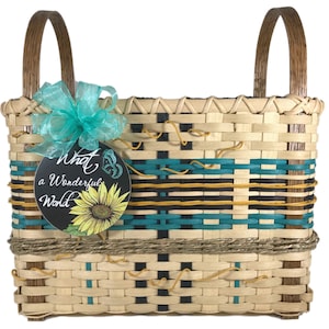 May include: A woven basket with a turquoise, black, and yellow pattern. The basket has two wooden handles and a turquoise bow. A tag with the text "What a Wonderful World" and a sunflower graphic is attached to the basket.
