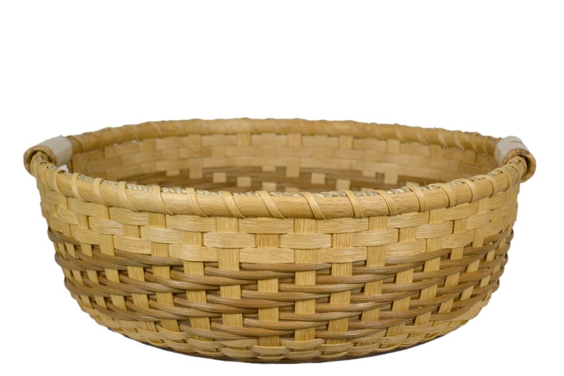 Pu&ograve; includere: Un cesto rotondo intrecciato con due manici in materiale naturale. Il cesto &egrave; di colore marrone chiaro.