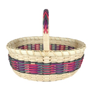 Puede incluir: Una cesta redonda tejida con una sola asa. La cesta está hecha de mimbre de color natural y tiene un diseño tejido colorido alrededor del borde superior en tonos de rosa, morado y verde.