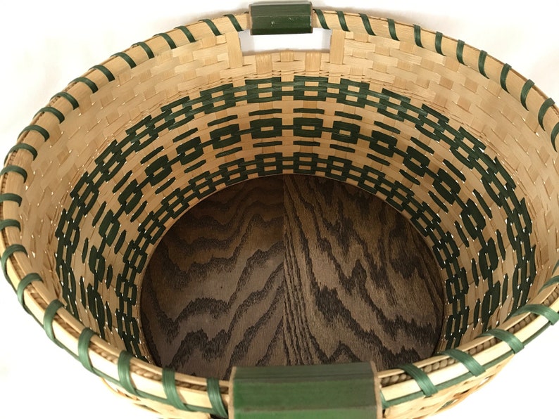 May include: A woven basket with a green and natural colored pattern. The basket has a wooden base and two green handles.