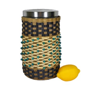 May include: A woven basket with a stainless steel lid and turquoise beads. The basket is brown and black with a woven pattern. A yellow lemon is sitting next to the basket.