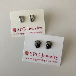 May include: Two pairs of silver skull stud earrings on white cards with the text "SPG Jewelry www.spgjewelry.etsy.com".
