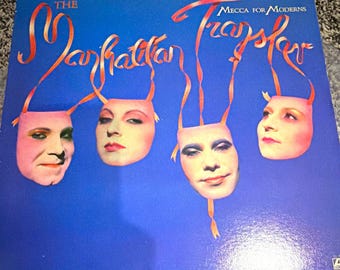 Op de afbeelding: Een vierkante vinylplatenhoes met een levendige blauwe achtergrond. De tekst luidt The Manhattan Transfer en Mecca for Moderns. Vier menselijke gezichten, afgebeeld als maskers, hangen aan rode linten tegen het blauwe veld.