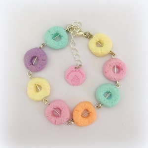 Puede incluir: Un brazalete colorido hecho de cuentas en forma de donut de colores pastel con una cadena plateada y un dije que dice "Sweets & Pearls".