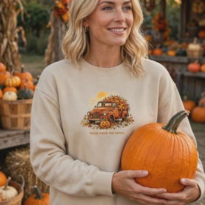 May include: A beige crewneck sweatshirt featuring a graphic of an orange vintage pickup truck loaded with pumpkins, surrounded by autumn leaves and a sun, with the text "FRESH FROM THE PATCH" printed below the truck illustration.