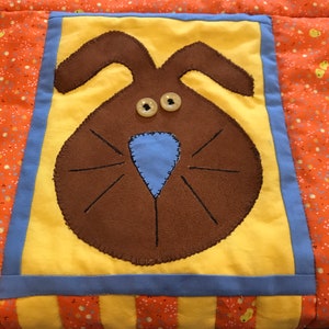 May include: A yellow and blue patchwork quilt square with a brown felt bunny face with blue nose and two yellow button eyes.