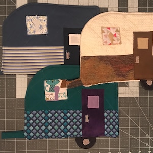 May include: Three quilted pot holders shaped like vintage campers. The first is blue with white stripes and a floral pattern. The second is beige with a brown door and a patchwork pattern. The third is green with a blue and white diamond pattern and a purple door.