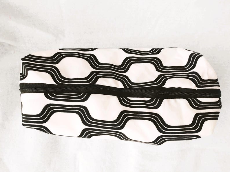 May include: A rectangular zippered pouch featuring a repeating black and white wavy geometric pattern. The fabric case has a black zipper closure running along the top center, with a soft, structured shape and a consistent graphic design.