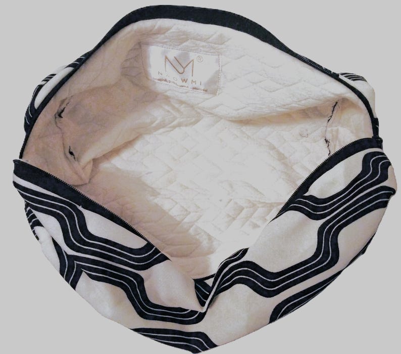 May include: A white and black zippered pouch with a wavy line pattern on the exterior. The interior is lined with a white quilted fabric featuring a rectangular brand label with the text NM and the word NAOWMI printed below it.