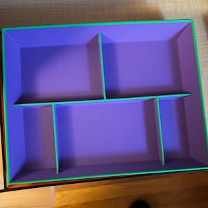 May include: A rectangular storage tray featuring a vibrant purple interior divided into four distinct compartments by bright green walls. The outer edges of the tray are also lined with a matching bright green border, creating a high-contrast look.
