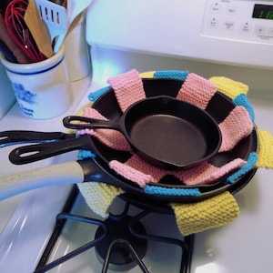 Puede incluir: Dos sartenes de hierro fundido anidadas, envueltas en un agarrador de olla de ganchillo de colores. El agarrador de olla está hecho de hilo rosa, azul y amarillo.