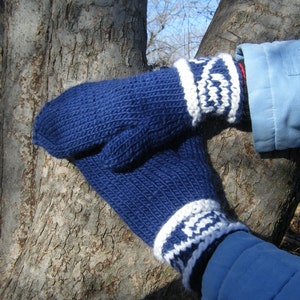 May include: A pair of blue knitted mittens with white trim. The mittens are worn by a person with a blue jacket.