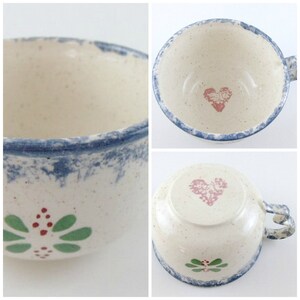 May include: A white ceramic mug with a blue rim and a pink heart design. The mug has a green and red floral design on the side.