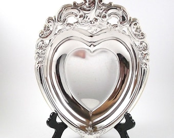 May include: Silver heart-shaped dish with an ornate, scrolled rim. The dish is on a black stand.