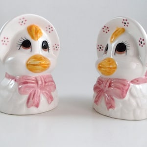 May include: Two white ceramic duck figurines. Each duck has a yellow beak, brown eyes, and a pink bow. They wear white bonnets with pink floral accents. The ducks have a vintage, whimsical design.