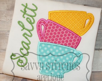 May include: A white t-shirt with the name "Scarlett" embroidered in green cursive lettering. The shirt features an applique of three stacked teacups in blue, pink and yellow with polka dot and checkered patterns. The text "Savvy Stitches Applique" is also embroidered on the shirt.