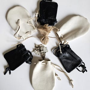 May include: Six small leather pouches, five are white and one is black, arranged in a circle on a white surface. A small bundle of dried sage is in the center of the circle.