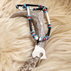 May include: A necklace featuring a white carved whale pendant suspended from a black cord. The strand consists of alternating long black and white cylindrical beads, interspersed with small round turquoise blue and red spherical beads.