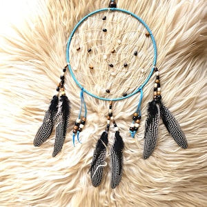 May include: A handmade dreamcatcher with a turquoise hoop and a detailed web. It has black and white speckled feathers, wooden beads, and small decorative beads. The dreamcatcher is designed for hanging and is set against a fluffy, cream-coloured background.