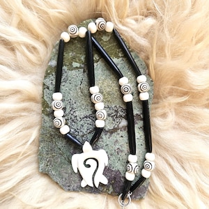 May include: A necklace featuring a white turtle-shaped pendant with a central spiral cutout. The strand consists of long, smooth black cylindrical beads alternating with smaller white spherical beads, some of which are decorated with black concentric circles.