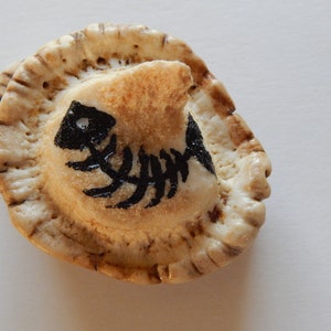 Puede incluir: Una pequeña costra de pastel de cerámica blanca con un diseño de esqueleto de pez negro pintado en la parte superior.
