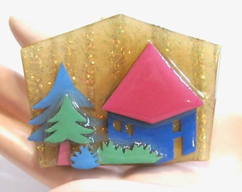May include: A vintage brooch featuring a blue house with a pink roof, a green tree, and blue and green bushes. The background is a glittery gold.
