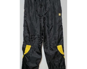 May include: Black nylon track pants featuring an elastic waistband with a drawstring, side pockets, and bright yellow accent panels on the lower legs. A small yellow Wilson logo is embroidered on the upper left thigh of the garment.