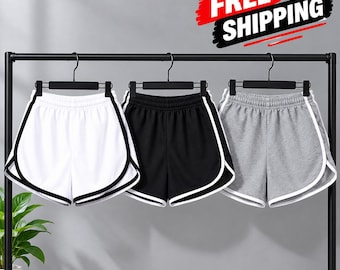 May include: Three pairs of athletic shorts hanging on black hangers. One pair is white with black trim, one is black with white trim, and one is heather grey with white trim. Each pair features an elastic waistband and curved hem.