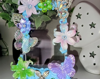 May include: A rectangular picture frame with a light blue textured border decorated with various colorful plastic flowers, iridescent butterfly figures, and clear rhinestone accents. The frame is mounted on a clear rectangular base.