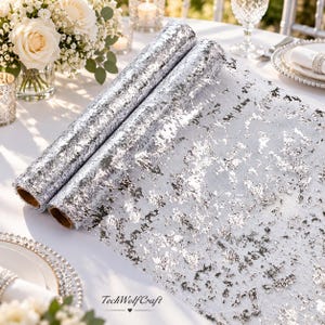 May include: A silver metallic foil table runner featuring a textured, crinkled finish with a reflective, shimmering surface. The runner is rolled into two cylindrical sections, displaying a pattern of irregular, shiny silver flakes.
