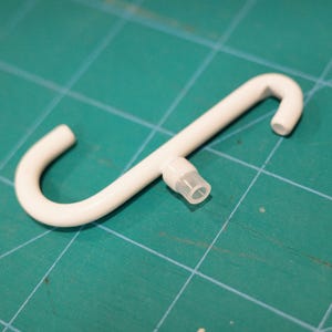May include: A white plastic S-shaped hook with a small, clear plastic tube extending from the center. The hook is on a green cutting mat.