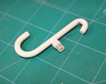 May include: A white plastic S-shaped hook with a small, clear plastic tube extending from the center. The hook is on a green cutting mat.