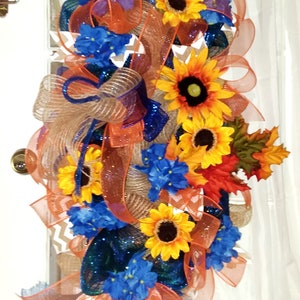 May include: A fall-themed door wreath with orange, blue, and brown mesh ribbon, sunflowers, blue flowers, and fall leaves.