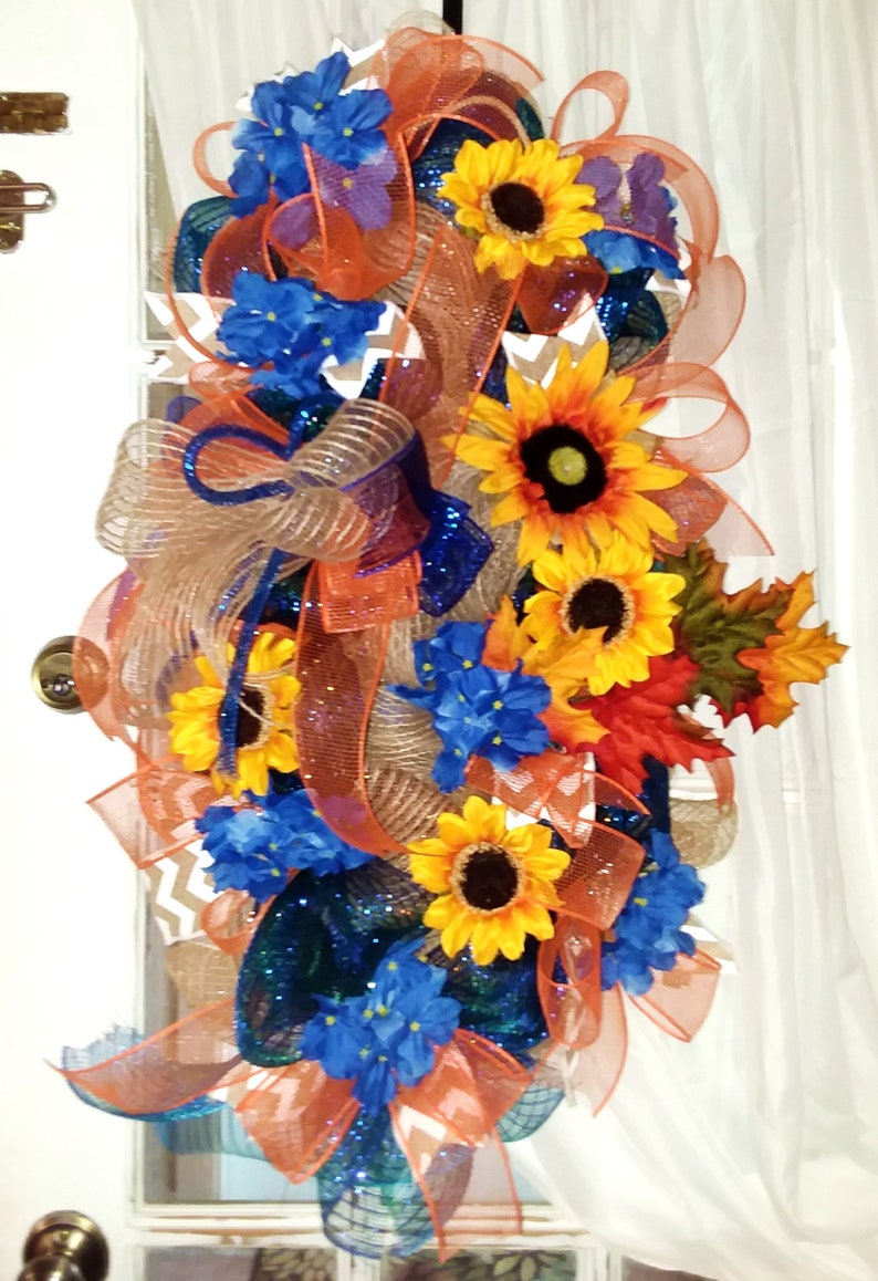 May include: A fall-themed door wreath with orange, blue, and brown mesh ribbon, sunflowers, blue flowers, and fall leaves.