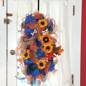 May include: A wreath with orange, blue, and yellow flowers, and brown and orange mesh ribbon. The wreath is hanging on a white door with a white sheer curtain.