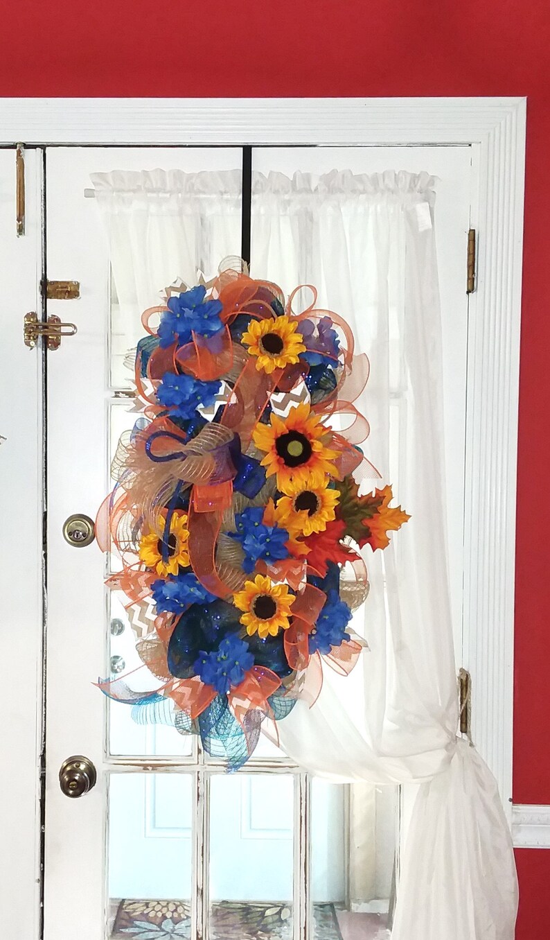 May include: A wreath with orange, blue, and yellow flowers, and brown and orange mesh ribbon. The wreath is hanging on a white door with a white sheer curtain.