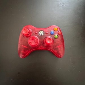 May include: A translucent red video game controller featuring a central Xbox logo button, two red analog sticks, a directional pad, and four colored action buttons labeled A, B, X, and Y, along with start and back buttons.