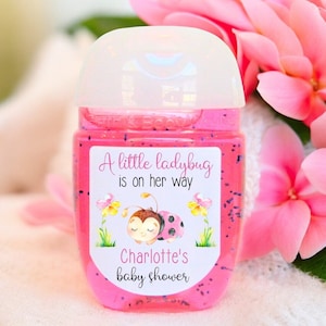 May include: Pink hand sanitizer bottle with a white label that says "A little ladybug is on her way Charlotte's baby shower" with a cartoon ladybug illustration.