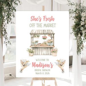 May include: A white sign on an easel with the text "She's Fresh Off The Market" in pink. Below is a watercolor illustration of a flower stand. The sign reads "Welcome to Madison's Bridal Shower March 8, 2026."