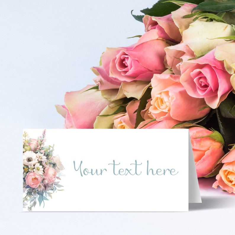 May include: A white card with a floral design on the left side and the text "Your text here" in blue script on the right side. The card is sitting on a light blue surface with a bouquet of pink roses in the background.